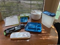 Assorted kitchen items on wooden table including mandolin, cutting boards, salad spinner, divided plates, veggie strip maker, cereal dispenser, and ice cube tray