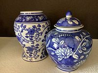 Two blue and white ceramic vases displayed side by side, one with lid and rounded knob, the other a baluster vase with floral scrollwork.