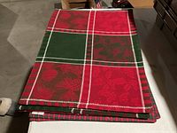 Red and green pinecone and berry patterned Christmas tablecloth folded and laying on the floor.