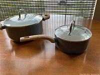 Two grey cooking pots with wooden handles and glass lids on a wooden table, one larger than the other.