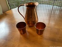 Set shows copper pitcher with curved handle and two matching copper glasses on wooden surface by window with blinds.