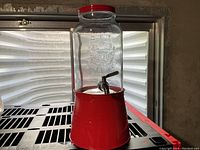 Glass water dispenser on the red stand, showing the spigot and embossed glass design