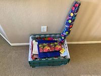 Photo shows plastic bin filled with various gift bags, partially unrolled roll of wrapping paper with balloon design standing next to bin in front of a beige wall.