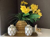 Photo showing two white ceramic vases with gold hexagonal pattern, a gold dog bookend, and a large textured gold vase holding yellow artificial flowers, all arranged on a gold colored rectangular tray.