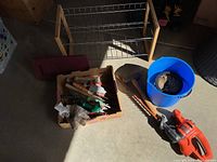 Photo showing gardening tools in a cardboard box, hammer, kneeling pad, blue bucket with soil, wooden shoe rack, and hedge trimmer on the floor