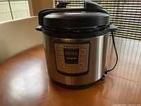 Front view of Instant Pot IP-LUX on wooden table showing stainless steel body and digital control panel with multiple preset buttons.