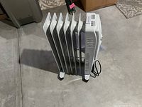 Side view of white seven-fin oil-filled radiator heater showing rotary temperature dial and power indicator light, caster wheels, carrying handle, and cord with manual attached.