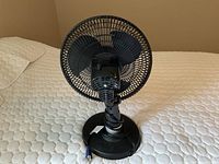 Front view of black Lasko oscillating fan showing blades and cage, placed on a bedspread.