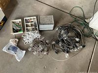 Two sets of outdoor hanging string lights with glass bulbs, wiring coiled on floor next to two white rope bundles and plastic containers with assorted screws and hardware.