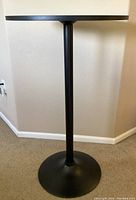 Side view showing the round black MDF tabletop and slender black metal cylindrical pedestal with flat wide base on carpet flooring.