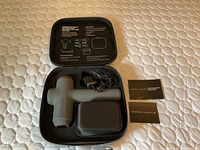 Top view of Sharper Image massage gun inside open carrying case with charging adapter and user manuals visible.