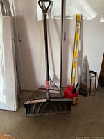 Snow shovel, leaf rake, saw, level, gas can and carpenter's square standing against a white cupboard, showing all items included in the lot.