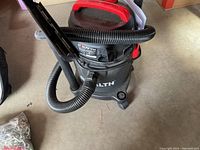 Black and red wet/dry vacuum with hose on concrete floor.