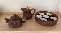 Set of two brown ceramic Asian teapots, six white-interior teacups, and circular serving dish with decorative carvings.