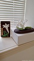 Bone china lily wall plaque and orchid flower sculpture on wooden base shown near window, displayed on white cardboard box.