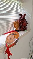Wide view showing large reddish-brown Buddha statue and attached oval wooden plaque with red string on windowsill.