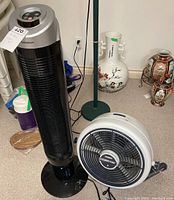 Black Gadason tower fan standing next to white Seabreeze Turbo-Aire turbo fan on carpet near ceramic vases.
