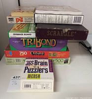 Stacked view of Scrabble, Pit, Cribbage, Trikond games and puzzle boxes showing titles and brands