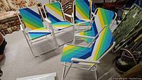 Group of five vintage lawn chairs with multi-colored diagonal striped cloth seats and backs and white metal frames, arranged indoors on carpet.