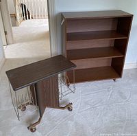 Photo showing both the vintage magazine rack/table with metal wire baskets and caster wheels and the wooden bookshelf with three shelves.