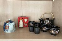 Photo shows a multicolor ceramic teapot with blue accents, a black ceramic teapot with floral design and woven handle, four matching black floral ceramic tea cups, and two small black sake cups.
