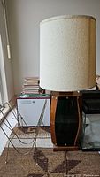 Photo showing vintage wooden lamp with cream-colored cylindrical fabric shade next to brass magazine rack on carpeted floor