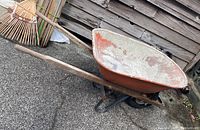 Side view of the wheelbarrow showing long wooden handles, metal tub with worn paint and single front wheel on concrete surface near wooden fence.