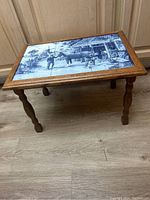 Full view of the wooden table with blue and white Delft-style tile top depicting the blacksmith shop scene.