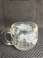 Clear glass mug with etched world map design, showing continents and grid lines, photographed against dark textured background.