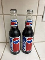 Two unopened Pepsi Longneck bottles with standard Pepsi labels, clear glass and metal caps, standing next to each other on a tiled surface.