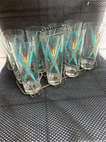 Group of 8 vintage tall glasses with turquoise and gold atomic starburst pattern arranged with metal caddy on a mesh background.