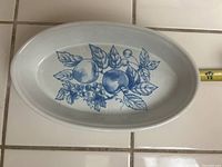 Top view of oval white ceramic bake casserole with blue fruit design and measuring tape for scale.