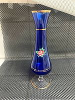 Front view of tall cobalt blue glass vase with hand painted pink rose and gold trim on woven black mat background
