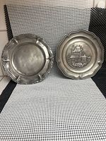 Two pewter wall hanging plates side by side on black and white checkered background, one plain and one embossed with Thun scene