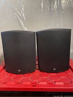 Front view showing two black BSI 870 monitor speakers with curved metal grills on red hexagonal plastic surface.