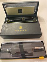 Two pens displayed in original Cross brand boxes: one black lacquer with gold trim, the other silver-tone in plastic tray.