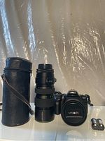 Full view of Minolta Maxxum 400si camera body with attached Hannimex 80-200mm zoom lens, leather lens case with strap, and two batteries placed next to camera.