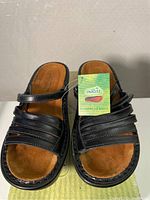 Front angled photo showing the left black leather sandal with cushion footbed and tag attached, labelled Naot.