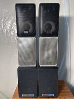 Photo shows six stereo speakers stacked vertically in pairs: at top two black Radio Shack speakers with fabric grills, in the middle two silver Realistic Minimus 7 speakers with metal grills showing dents, bottom two black Koss speakers with fabric grills.