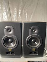 Pair of black active studio monitors with visible tweeter, woofer, and bass port against a white background surface and a clear plastic sheet in the backdrop.