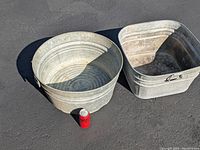Photo showing one square galvanized steel wash tub alongside one round galvanized tub, with soda can placed next to both tubs for size reference, on asphalt ground.