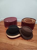 Two men's medium antique hats on the floor, one black and one brown, next to open burgundy oval hat boxes with beige interiors.