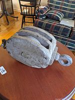 Large wooden sailboat pulley, side view showing bulkiness and thickness of wooden frame, placed on round wooden table indoors near plaid upholstered chair.