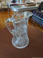 Full view of the clear cut glass crystal pitcher showing detailed cut patterns and flared lip on wooden table with living room setting in background