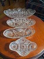 Four clear crystal trays arranged on a wooden table showing detailed cut designs and shapes, including oval and scalloped edges.