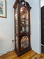 Front view of tall lighted wooden display cabinet with glass shelves and doors, showing the intricate wood curved top and bottom design.