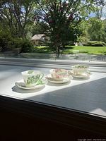 Three festive teacups and saucers displaying floral designs placed on a windowsill with outdoor greenery visible in background.