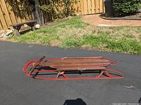 Side view of a Flexible Flyer sled showing wooden slats seat and red metal runners on pavement outdoors.