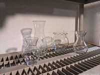 Four clear glass flower vases shown on a shelf with visible details of shapes and sizes