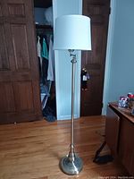 Photo of a floor lamp standing on a hardwood floor in a room with wooden closet doors and some furniture in the background.